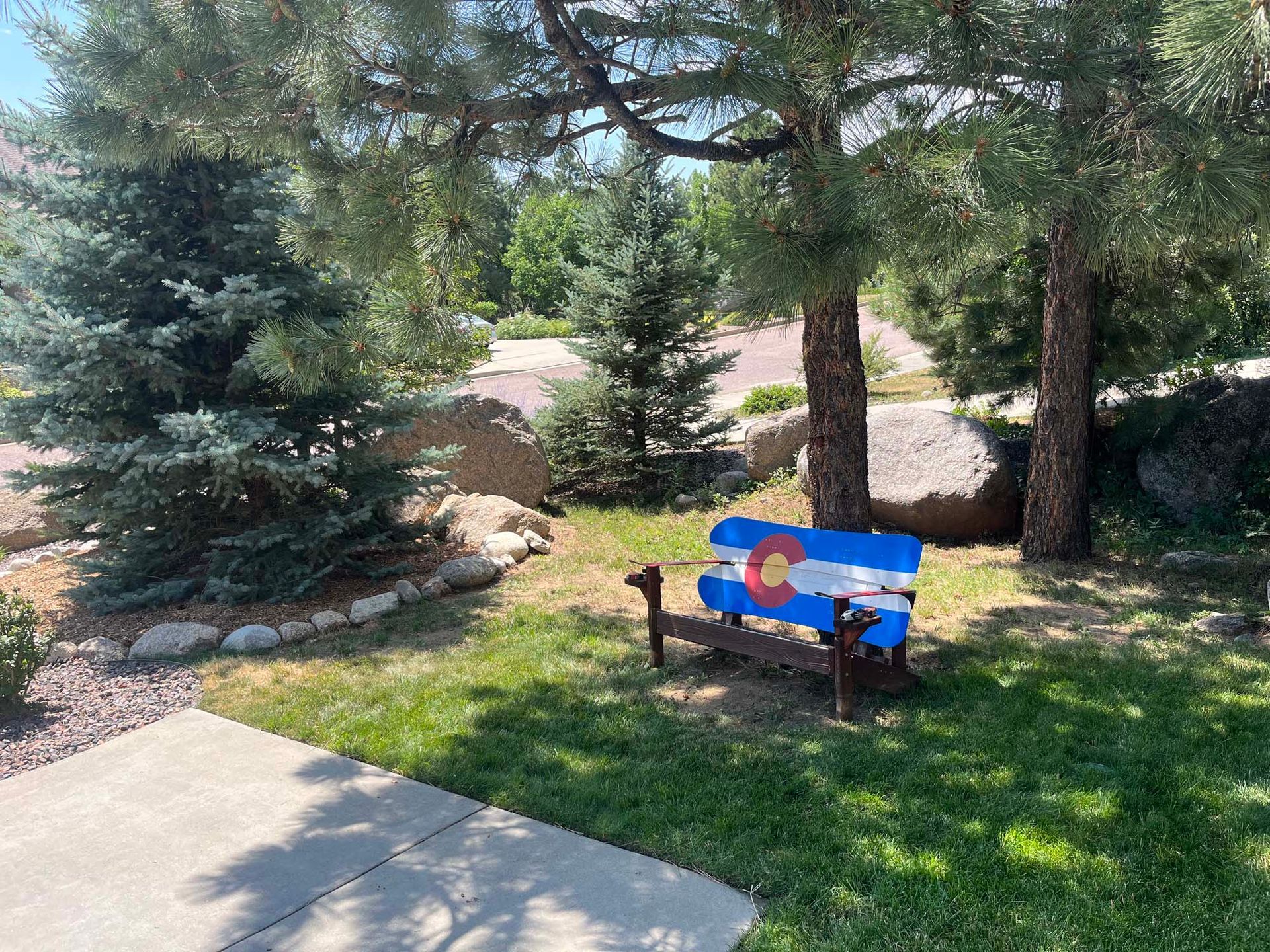 Bench painted with the Colorado flag in a grassy yard, under trees.
