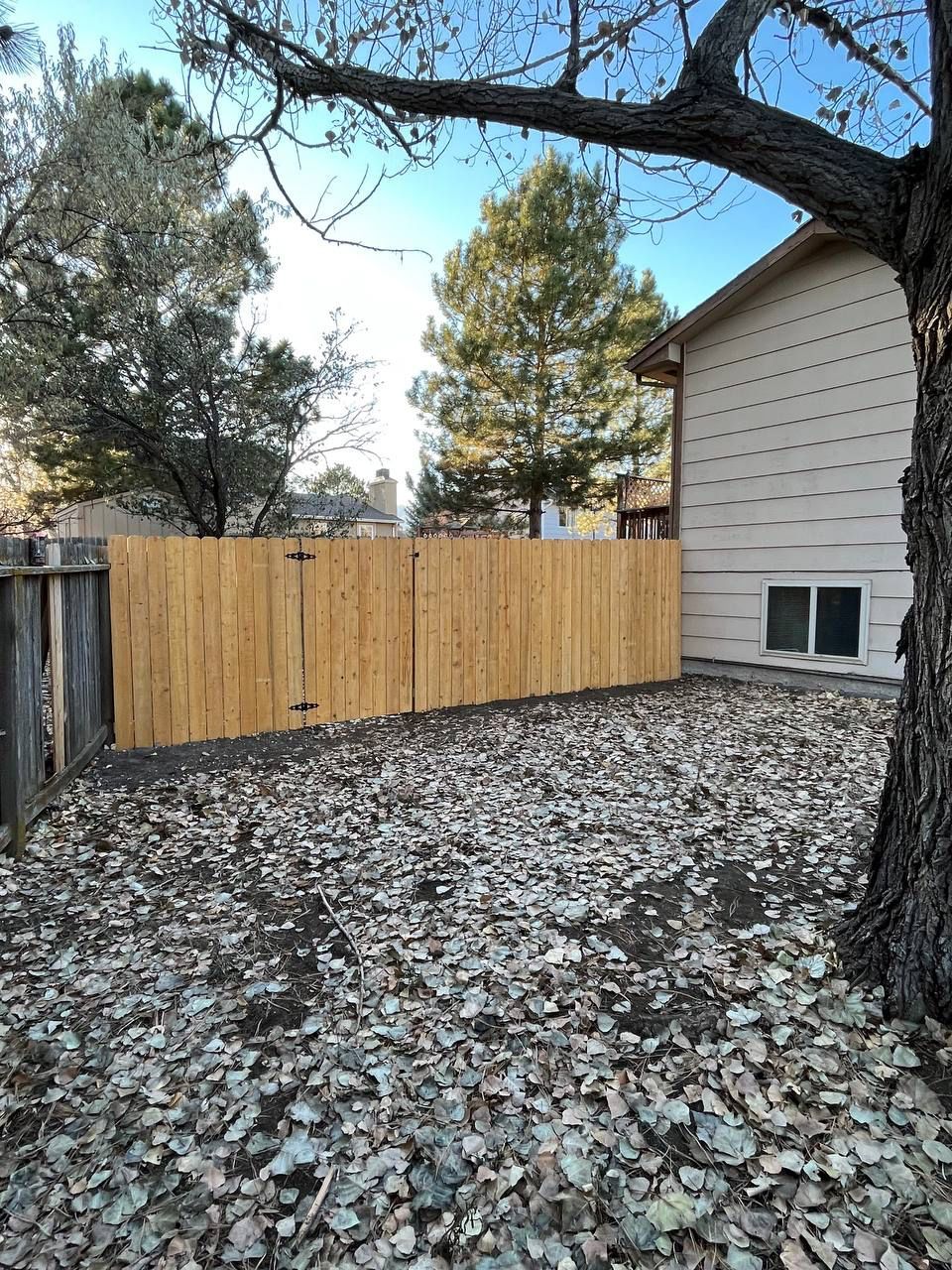 A wooden fence is in the backyard of a house surrounded by leaves.