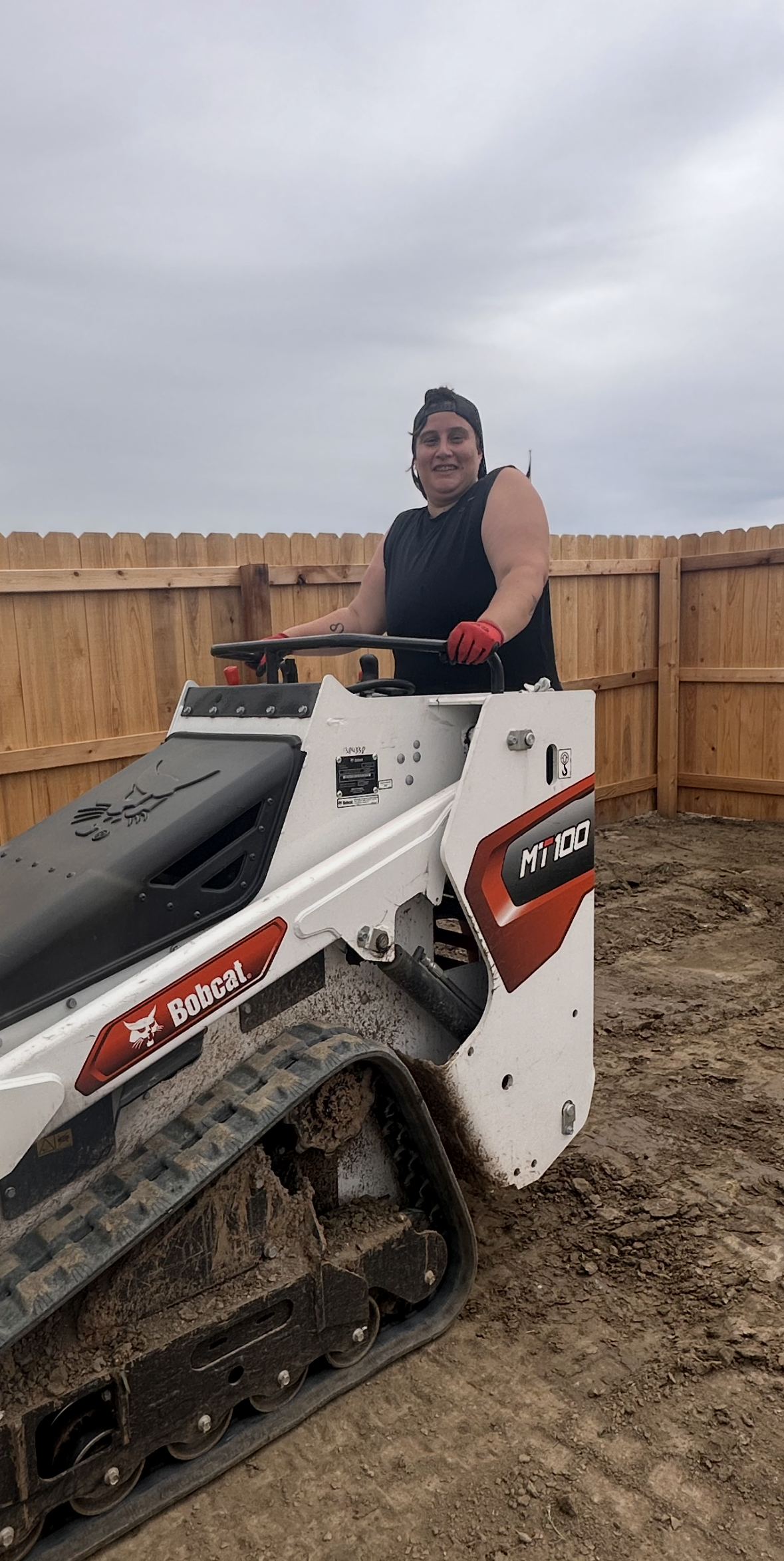 Person operating a Bobcat compact track loader in a yard, smiling. Cloudy sky and wooden fence in the background.