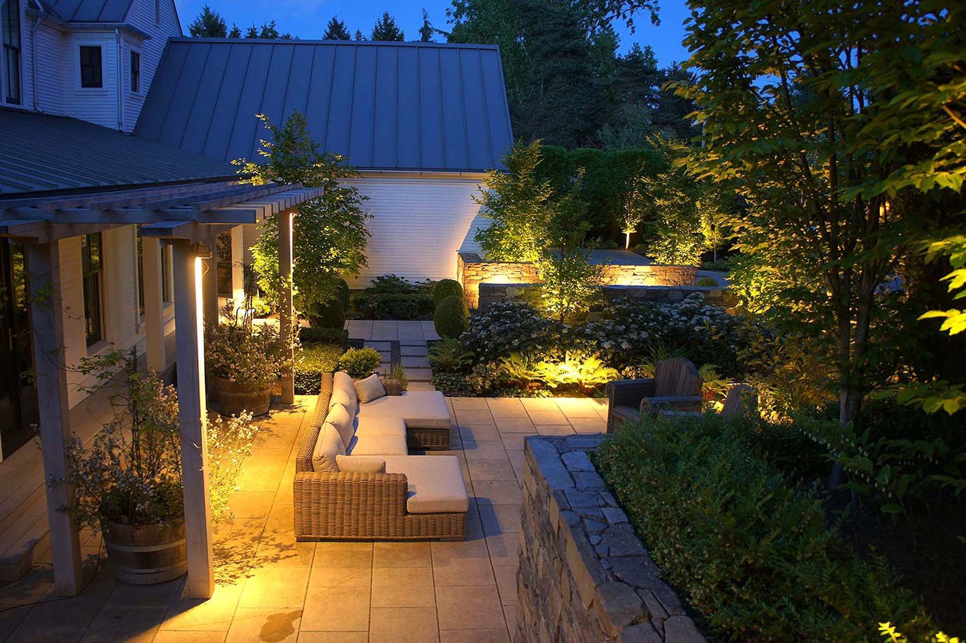 Nighttime outdoor patio with warm lighting illuminating a stone walkway, seating area, and landscaping.