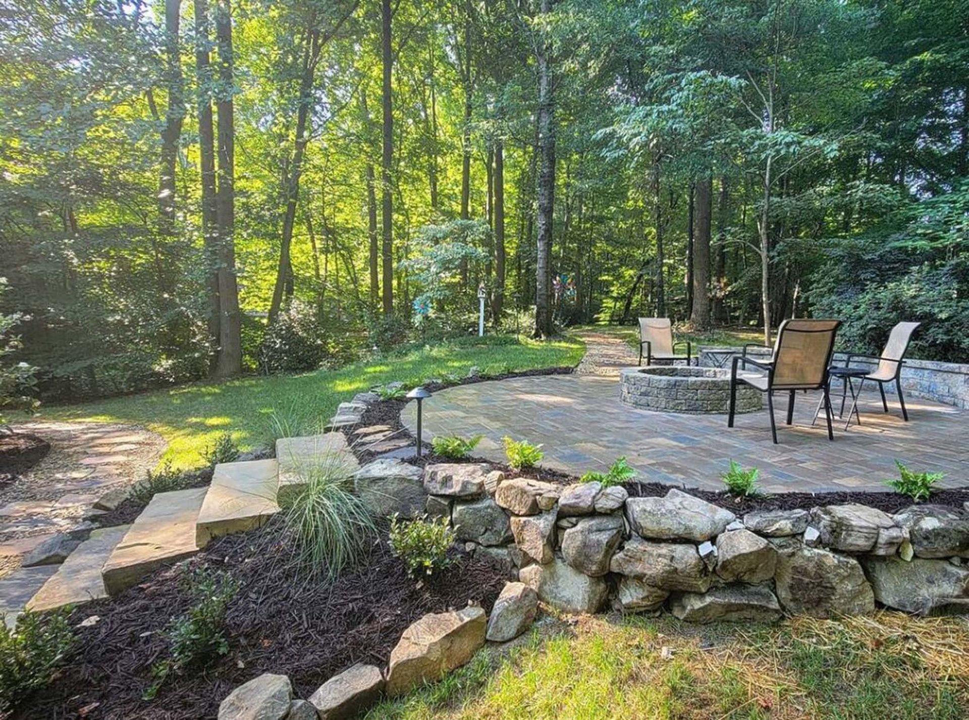 Stone patio with fire pit, seating, and stacked stone retaining wall in a wooded backyard.