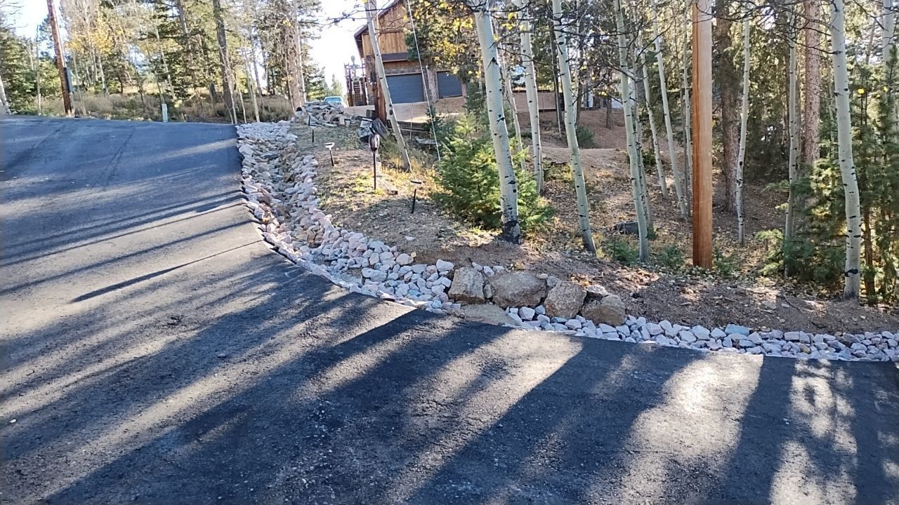Asphalt driveway with rock border, leading to a house in a wooded area.