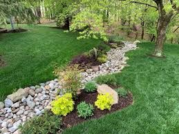 A dry creek bed winds through a green lawn, surrounded by colorful plants and a tree.