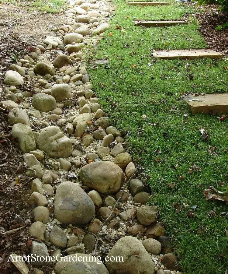 Stone-lined pathway with rocks and gravel next to a grassy lawn with stepping stones.