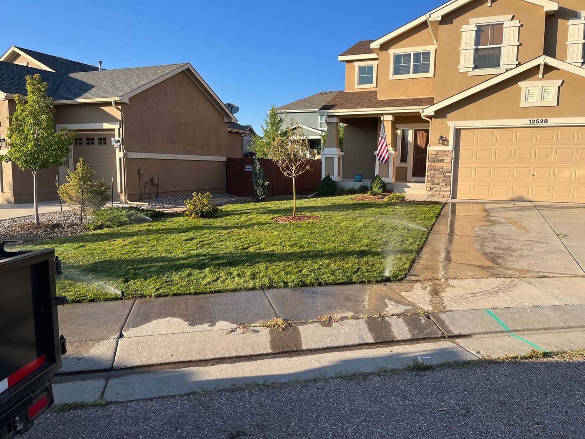 Lawn sprinkler watering grass in front of a beige two-story house with a brown roof and a smaller house to the left.