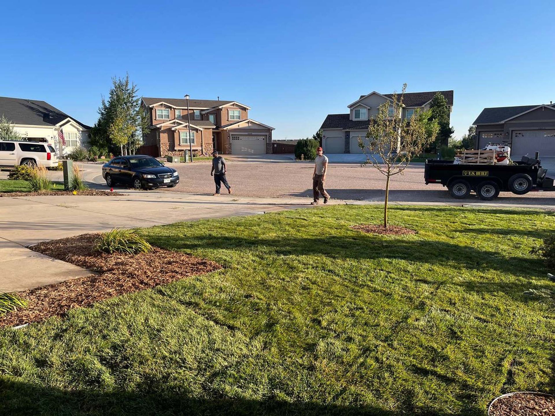 Two people walking on a street, a black car, a trailer, and houses under a clear blue sky.