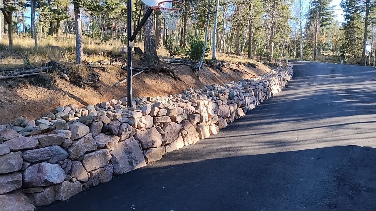 A basketball hoop is sitting on the side of a road next to a stone wall.