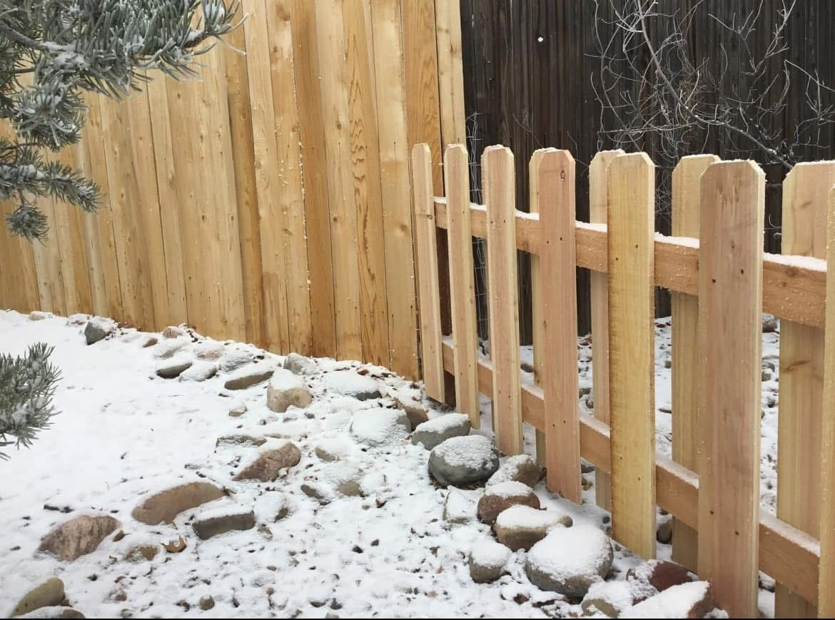 A wooden picket fence is surrounded by snow and rocks
