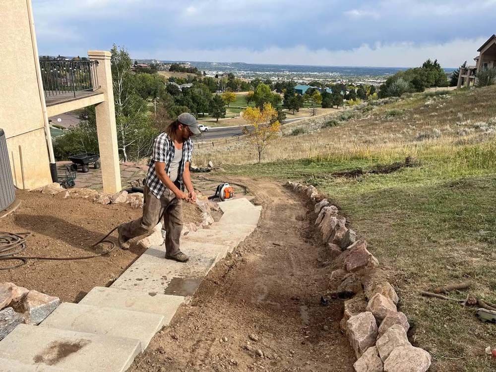 Man using tool on path next to a house with a scenic hillside view.