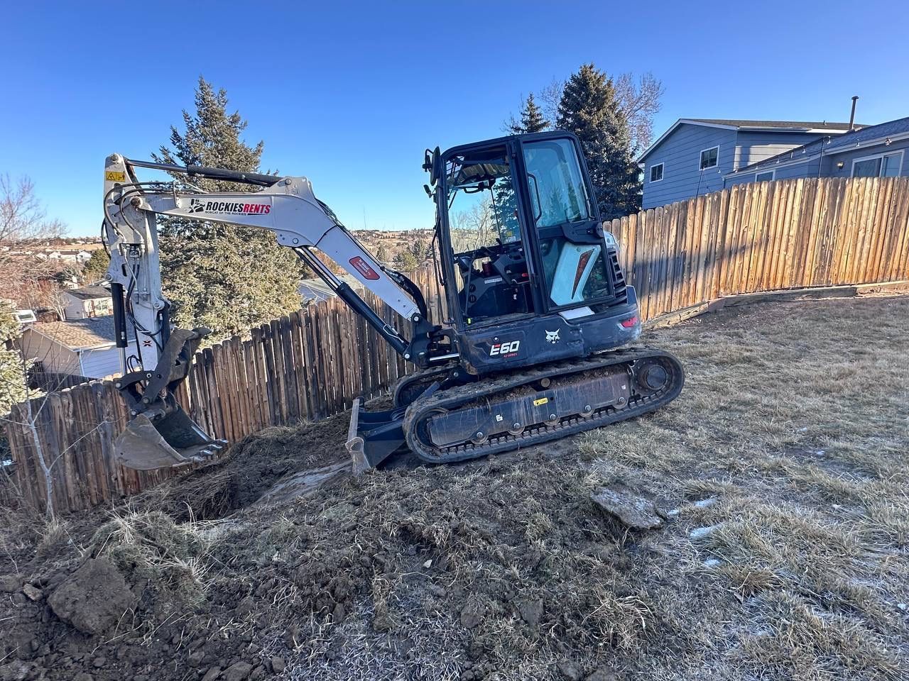 A small excavator is digging a hole in the dirt in front of a fence.
