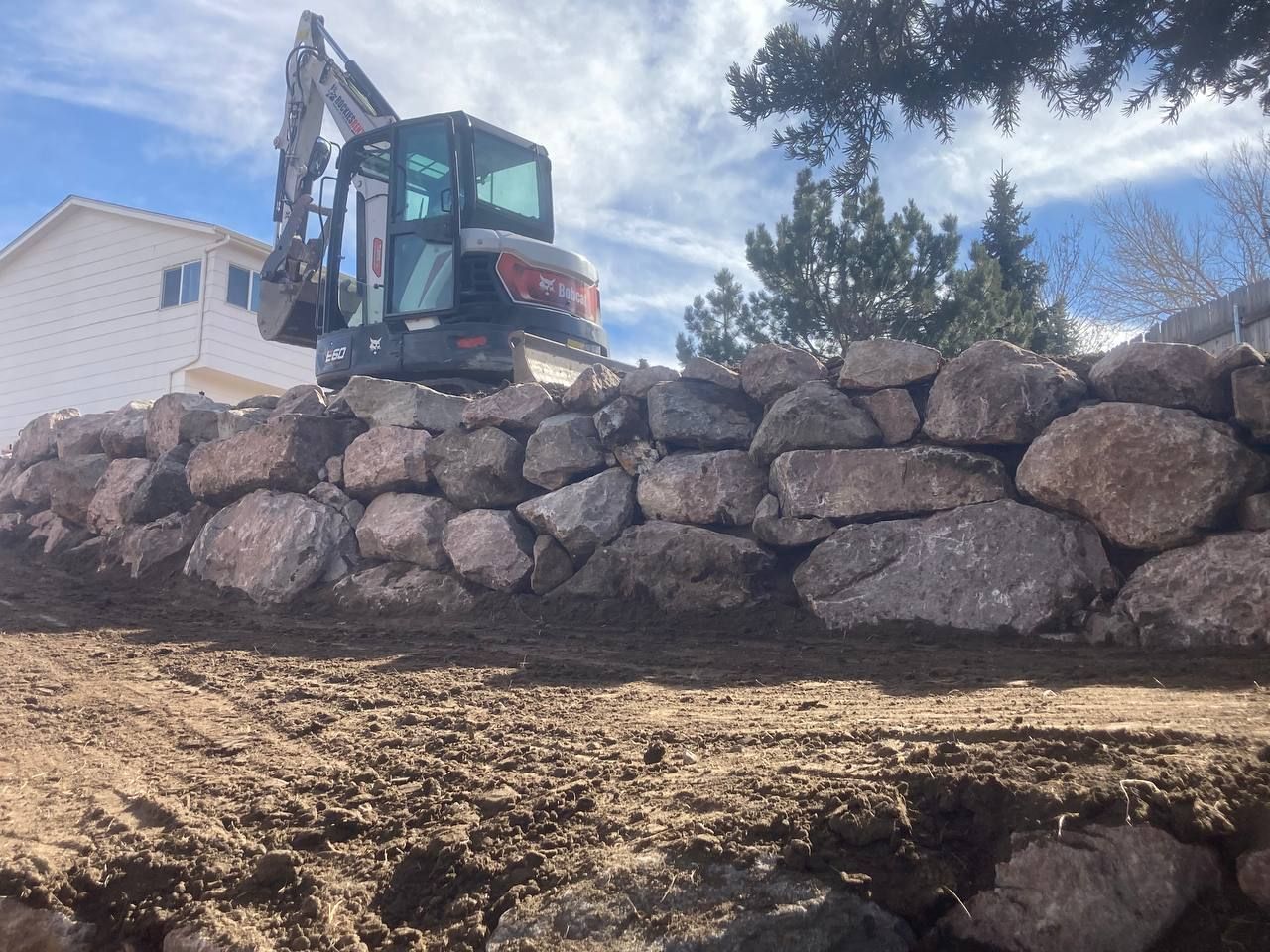 An excavator is working on a rock wall in front of a house