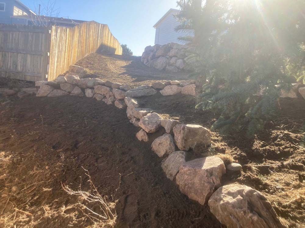 Stone retaining wall on a hillside with a wooden fence and house in the background.