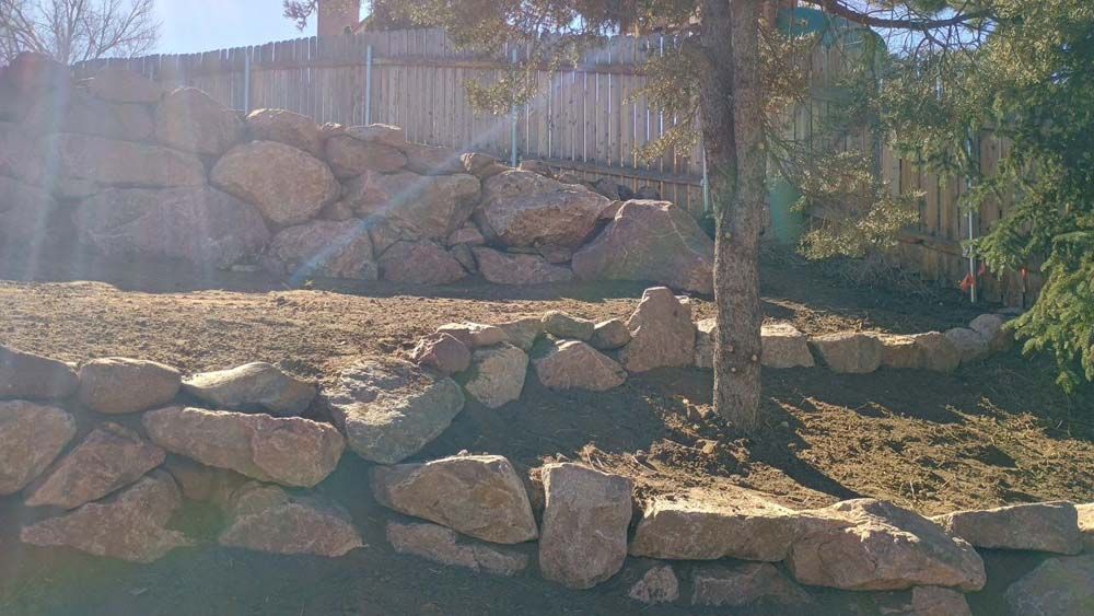 Rocky retaining walls and sloped ground in a sunny outdoor setting with a tree and a wooden fence.