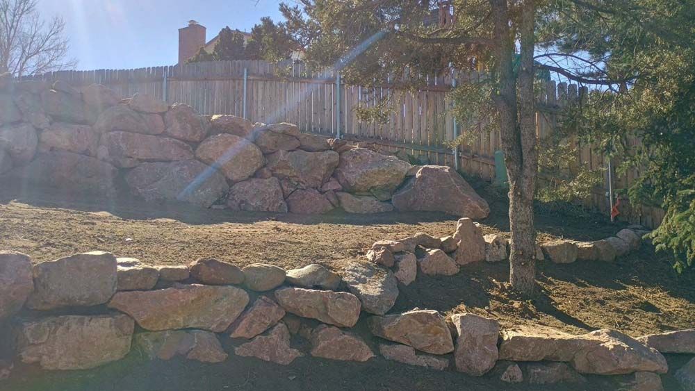 Rock retaining walls on a sloped yard with a fence and tree.