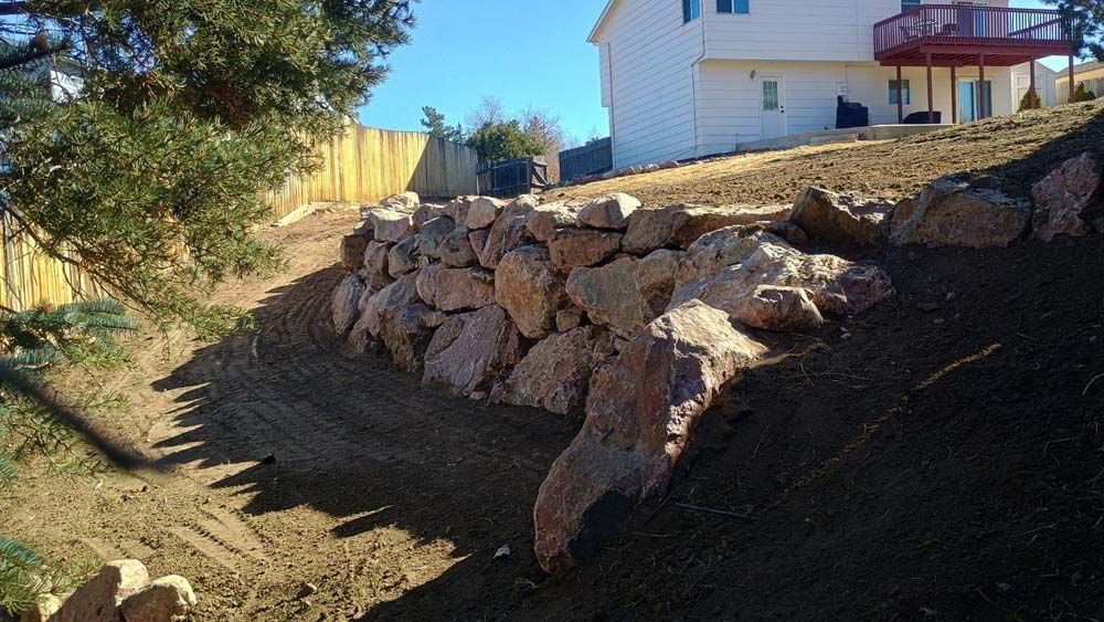 Rock retaining wall on a hillside with house in the background and a fence.