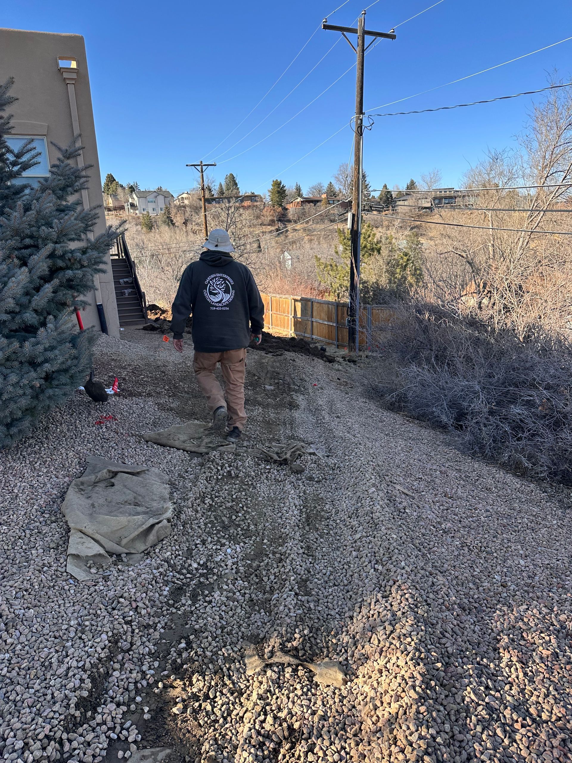 A man wearing a black jacket with a logo on the back walks through a gravel area
