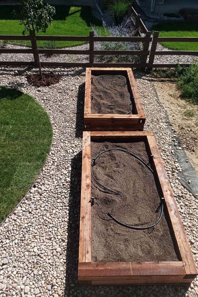 Two raised wooden garden beds filled with soil, one with irrigation tubing, in a gravel yard.