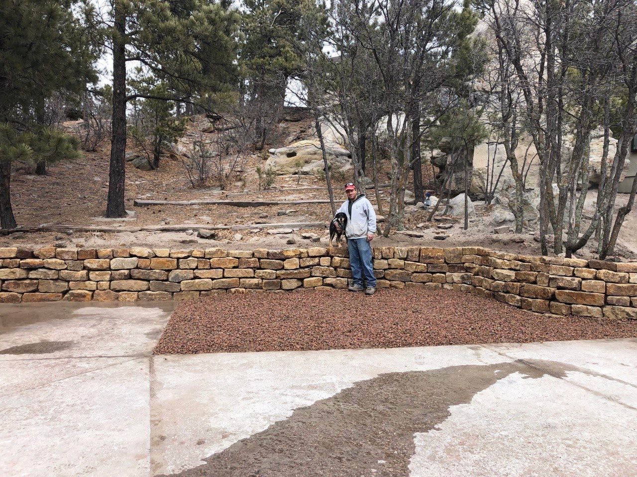 A man is standing in front of a stone wall in the woods.