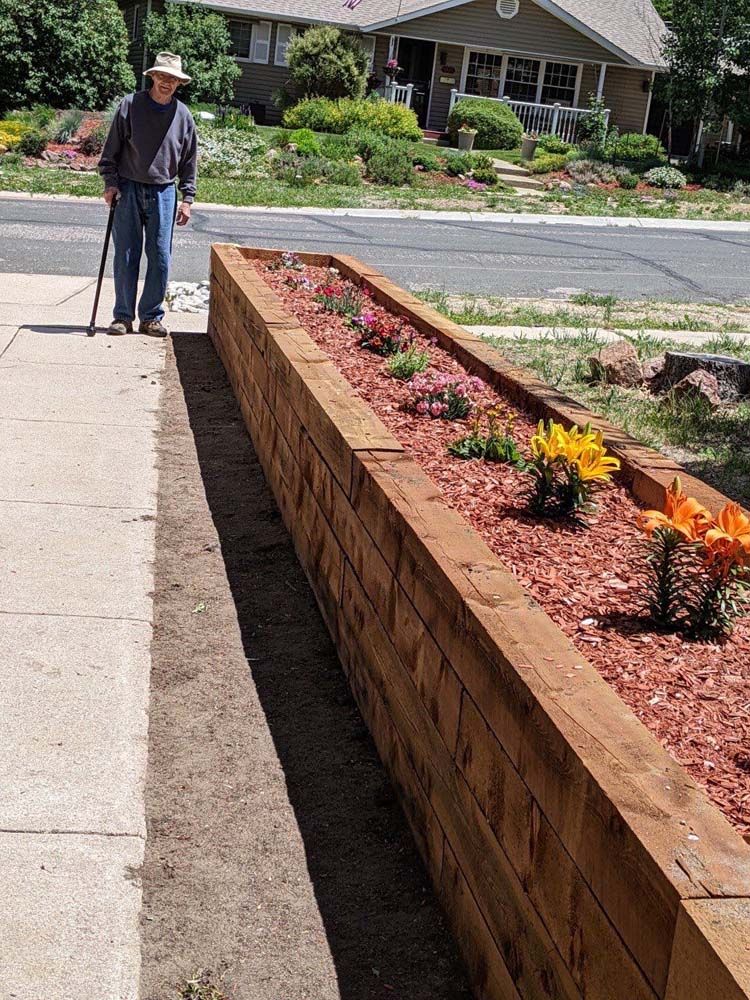 Man with cane stands next to a wooden planter box filled with flowers on a sidewalk near a house.