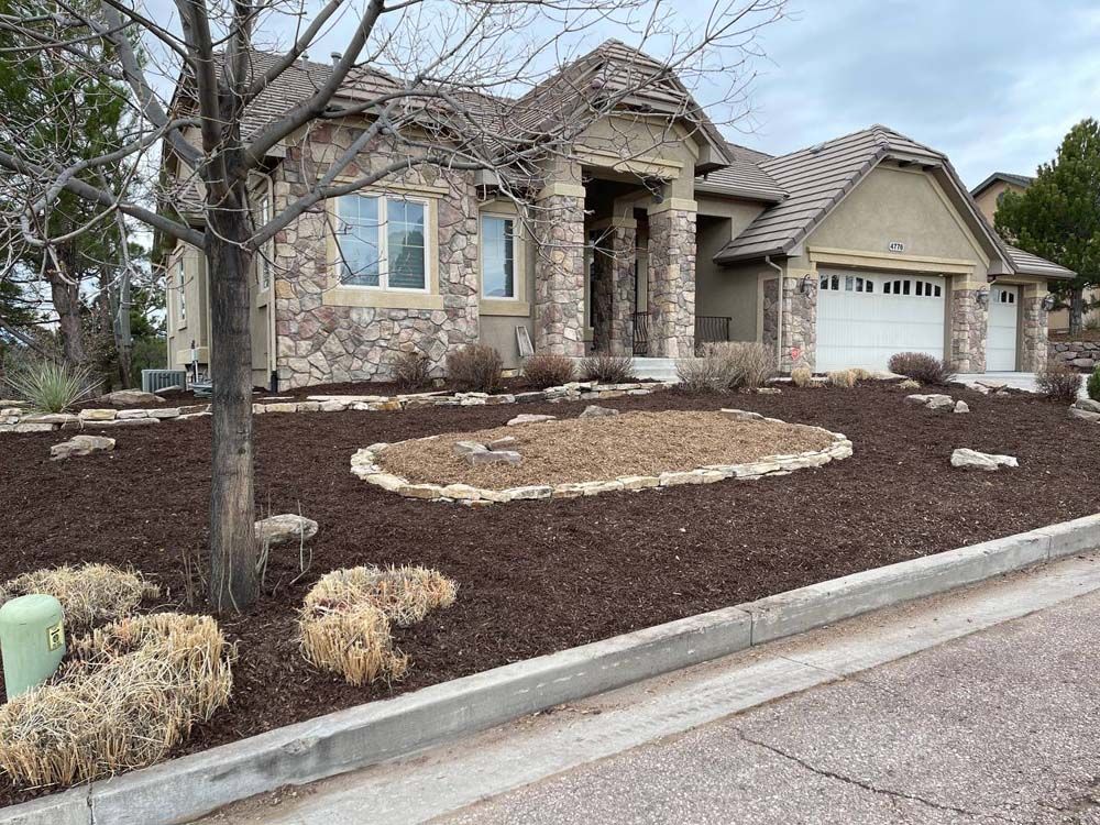 Stone-faced house with brown mulch landscaping and a dry, brown lawn. Gray sky.