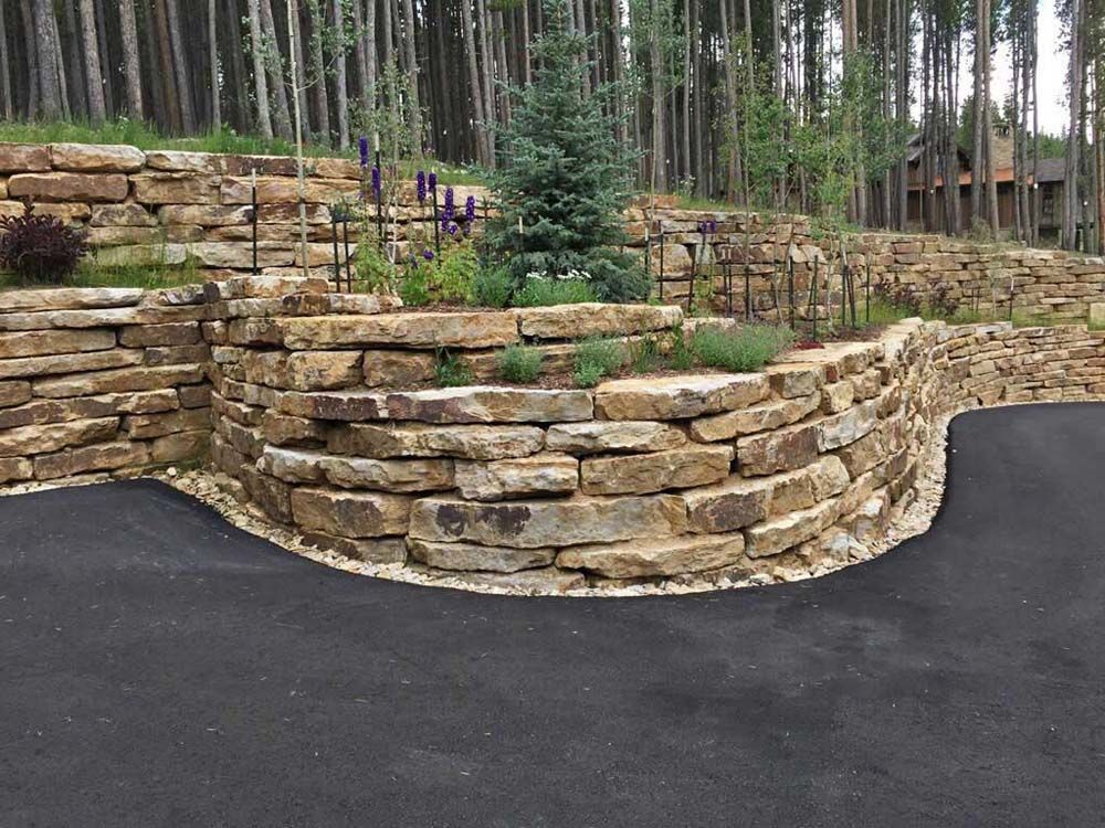 Stone retaining wall with plants, bordering an asphalt driveway, and forest in background.