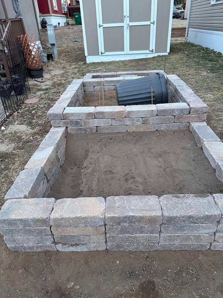 Two rectangular raised garden beds made of gray bricks, on dirt ground. A shed is in the background.