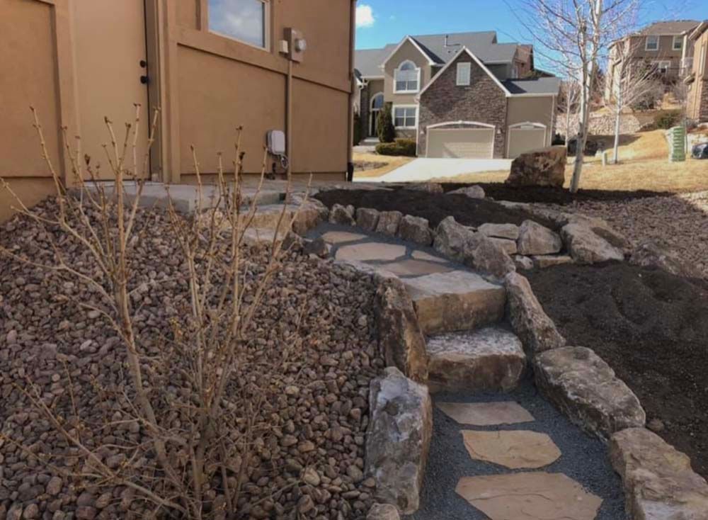 Stone pathway with steps, bordered by rocks and gravel, leading to a beige building, sunny day.