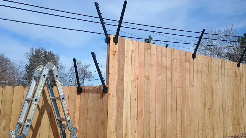 Wooden fence with black angled extensions, ladder leaning against it, power lines above.