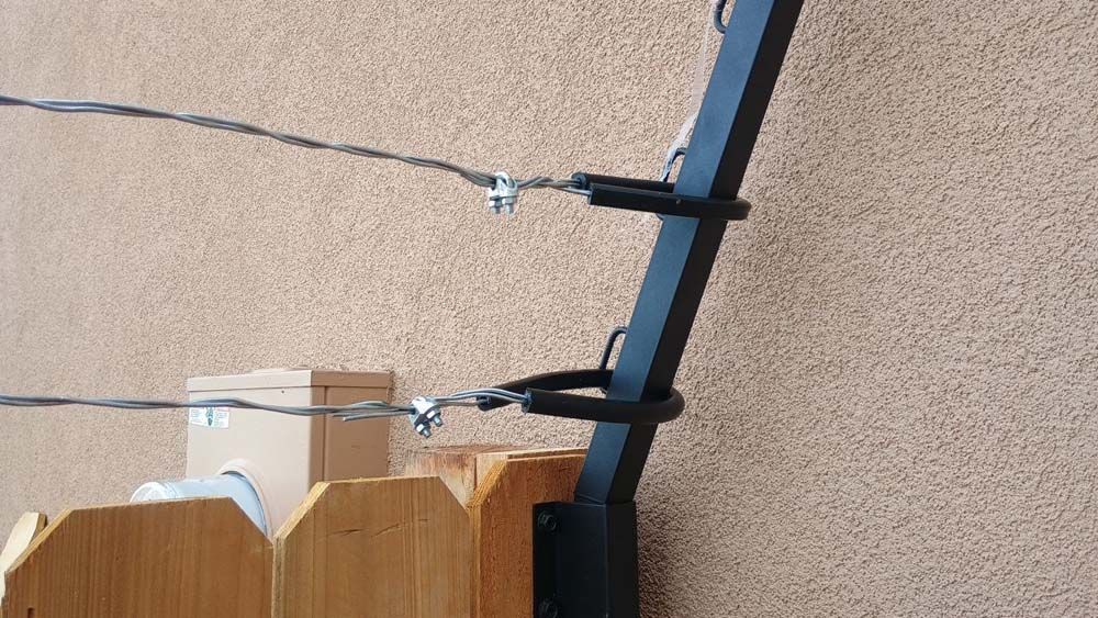 Black metal post with wires attached by metal clips; wooden fence in foreground, tan stucco wall in background.