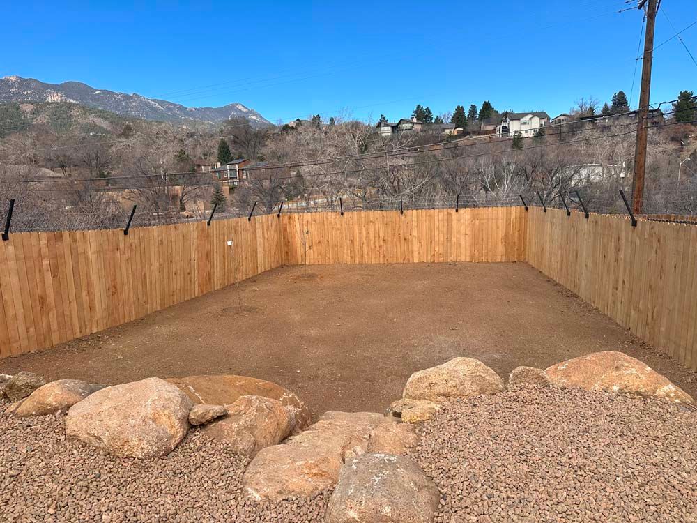A fenced-in yard with rocks, dirt, and a mountain backdrop under a clear blue sky.