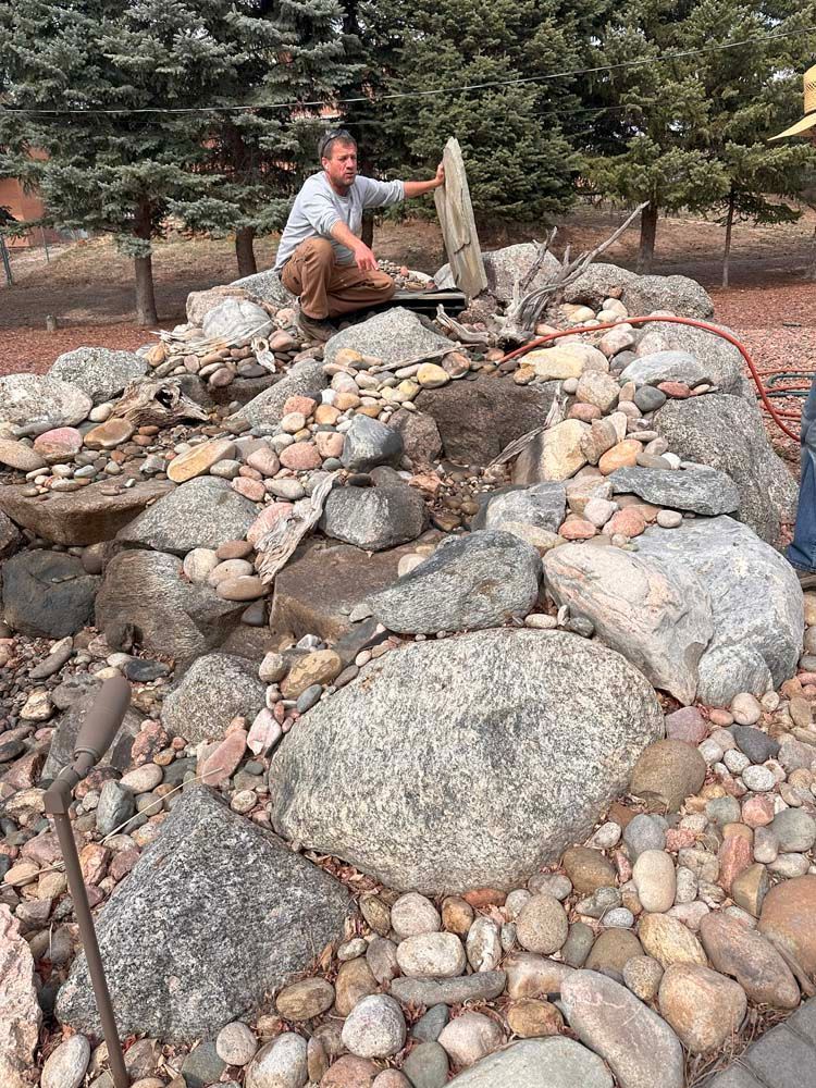 Man adjusts a wooden lid on a rock waterfall. He's outdoors among rocks and trees.