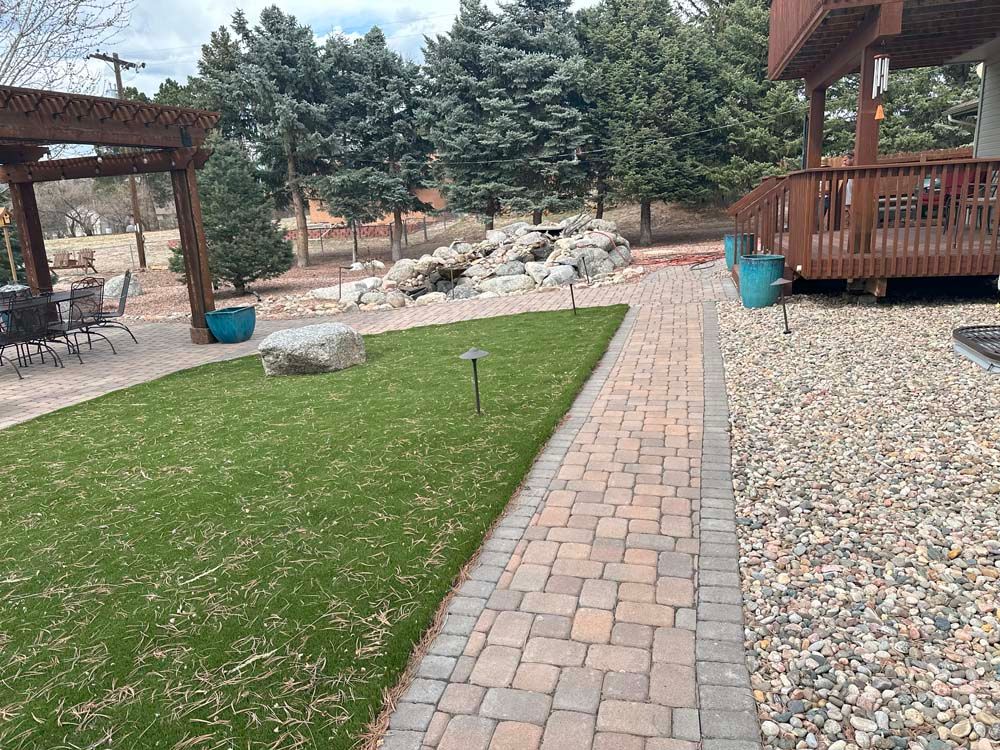 Brick pathway and green lawn lead to a deck and pergola in a backyard.