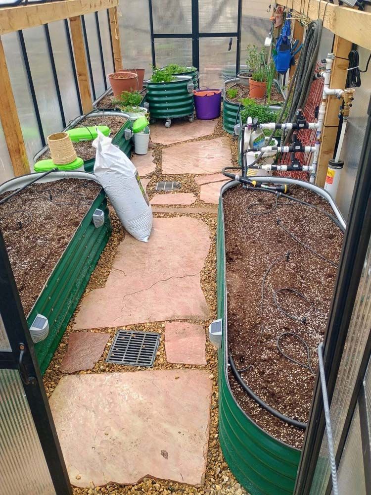 Greenhouse interior with stone path, raised garden beds, and various potted plants.