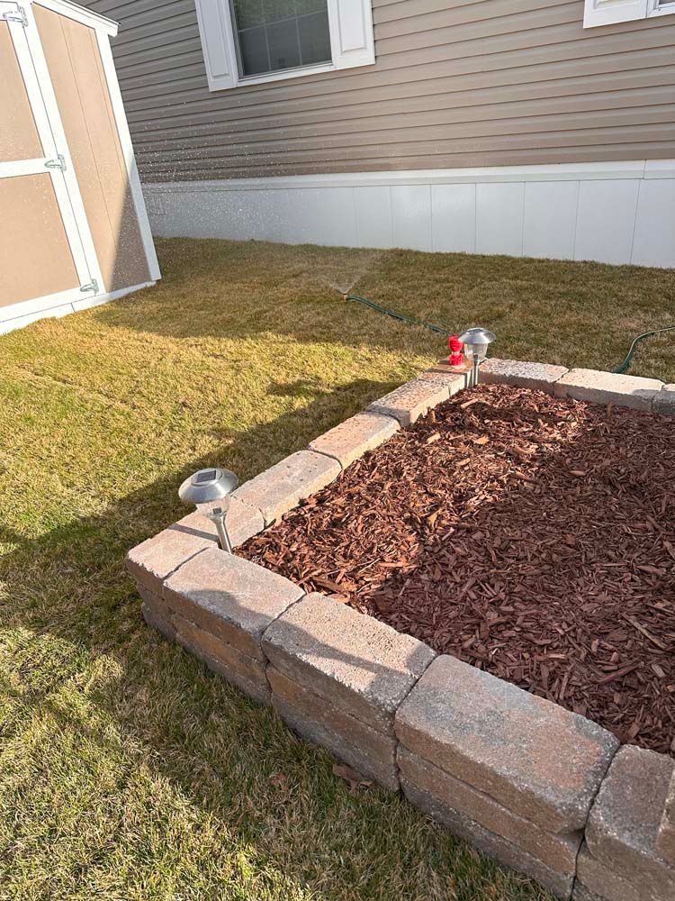 Raised garden bed with brown mulch and brick edging, solar lights, and a shed on a lawn.