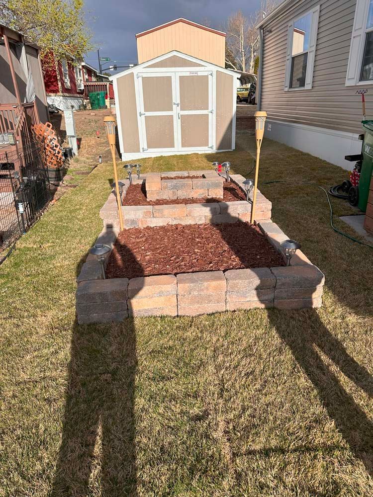 A brick fire pit with decorative torches and red mulch in a backyard setting.