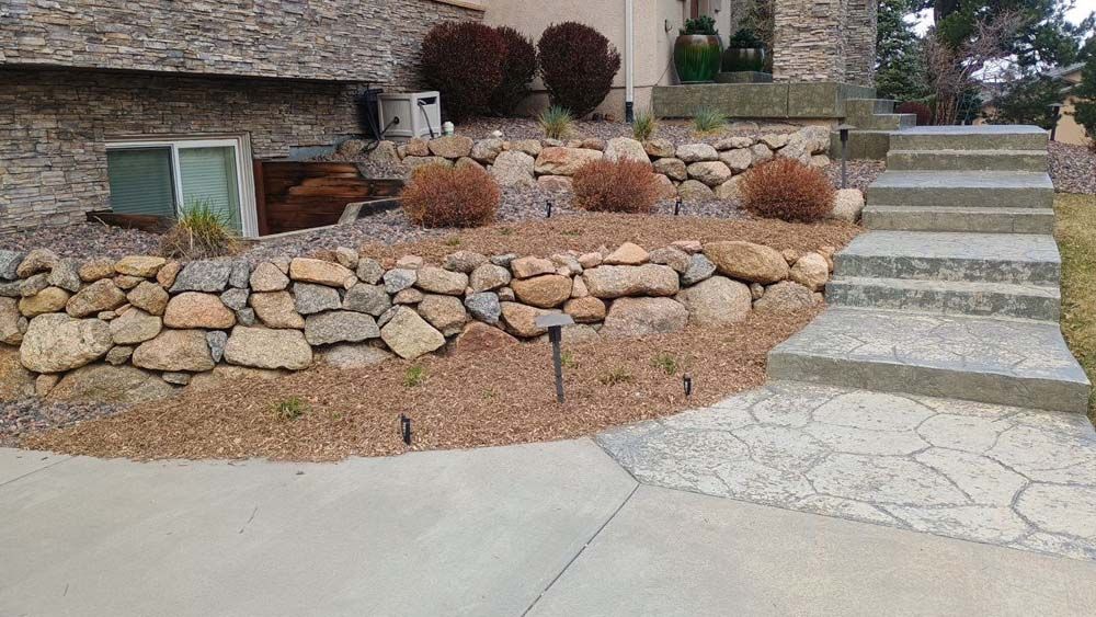 Stone retaining wall with steps leading up to a house. Brown mulch and dry bushes are in the landscaped area.