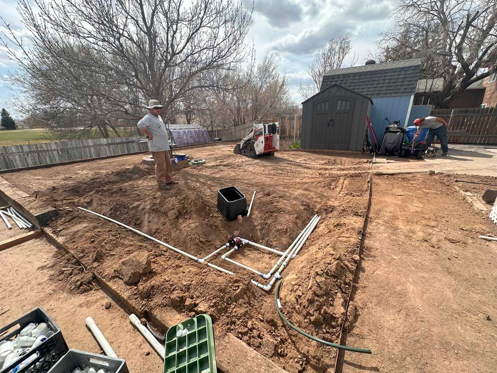 Construction site with workers, machinery, and plumbing, digging in dirt, near a house and trees.