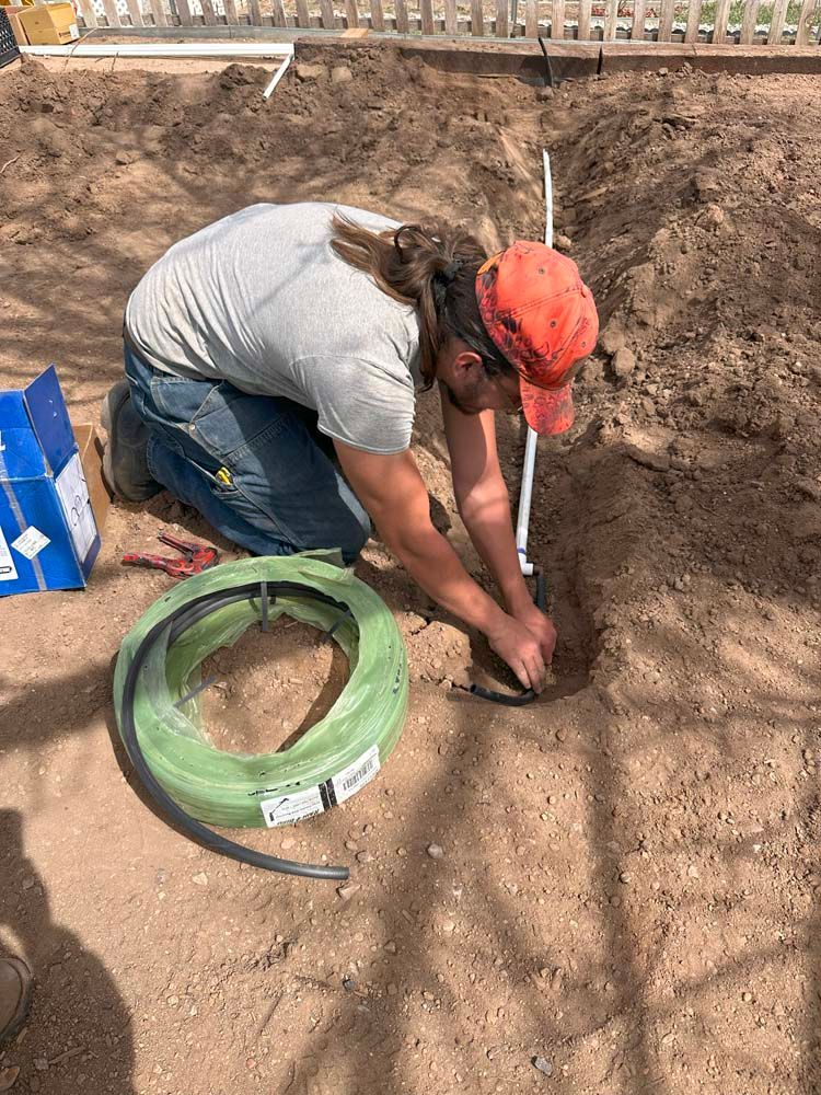 A person in an orange cap kneels, installing a black hose into a hole in the dirt; irrigation work.