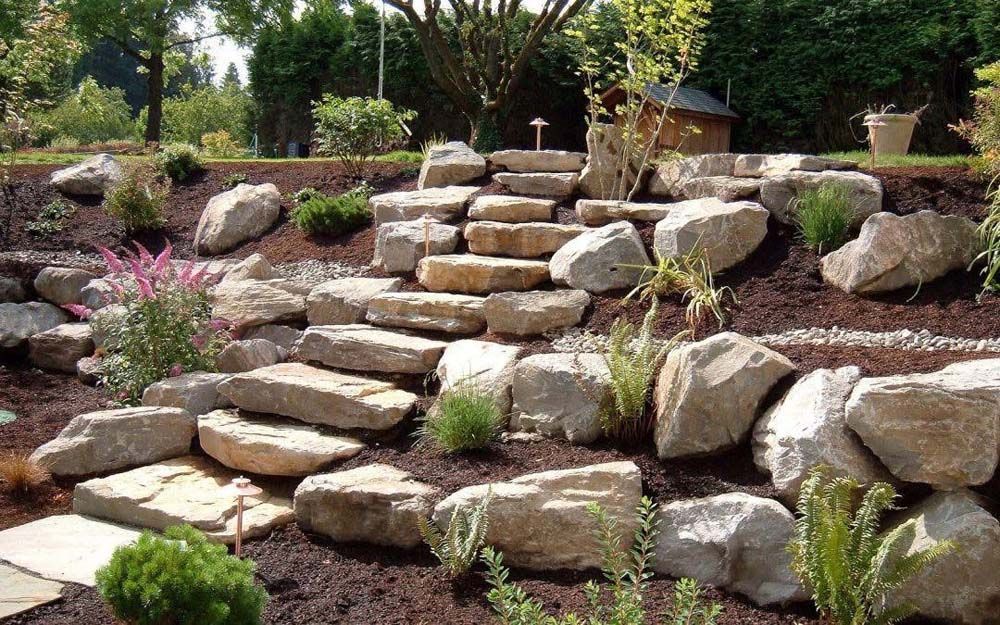 Stone steps ascend a sloped garden bed filled with plants and large rocks.