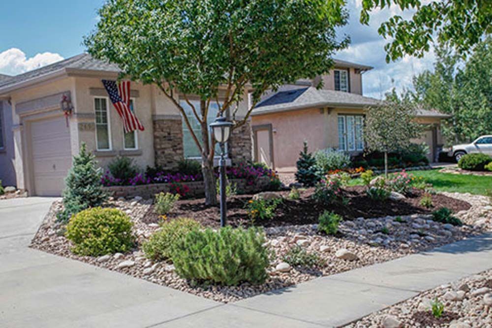 Tan house with landscaped front yard, American flag, trees, shrubs, and stone edging.
