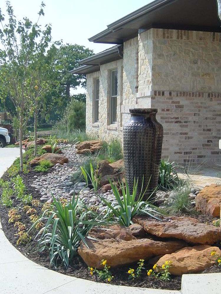 Landscaped yard with stone rocks, water feature, and plants next to a brick house.