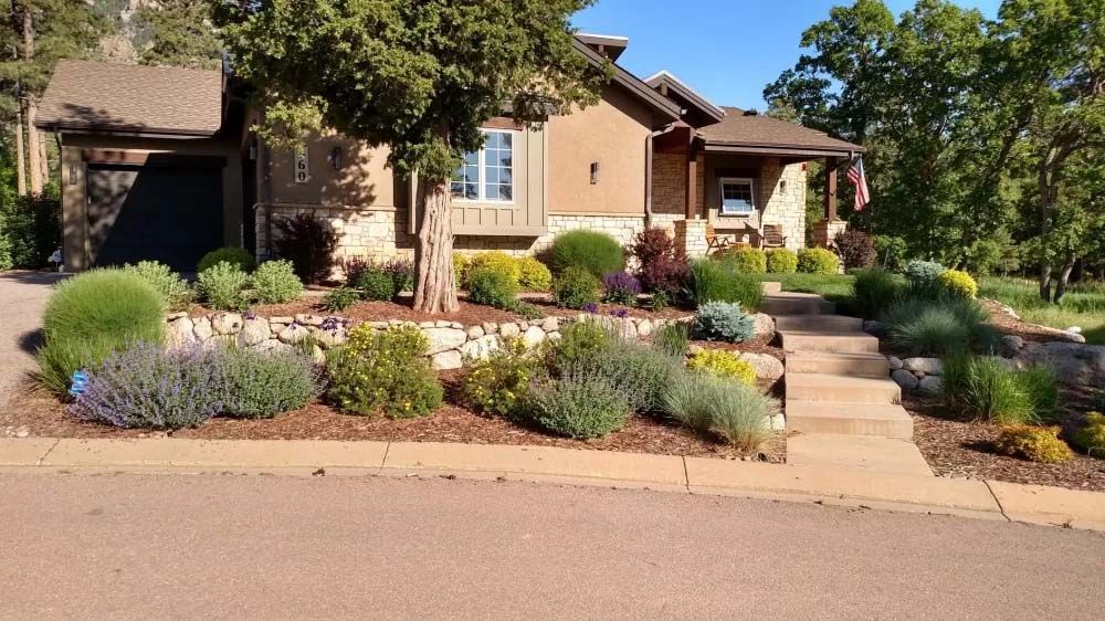 House with garden beds, stone walls, and a pathway leading to the front door. Lush greenery and blue sky.