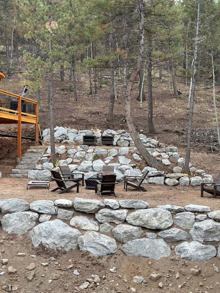 Stone terraced outdoor seating area with chairs, a grill, and a wooden deck in a forest setting.