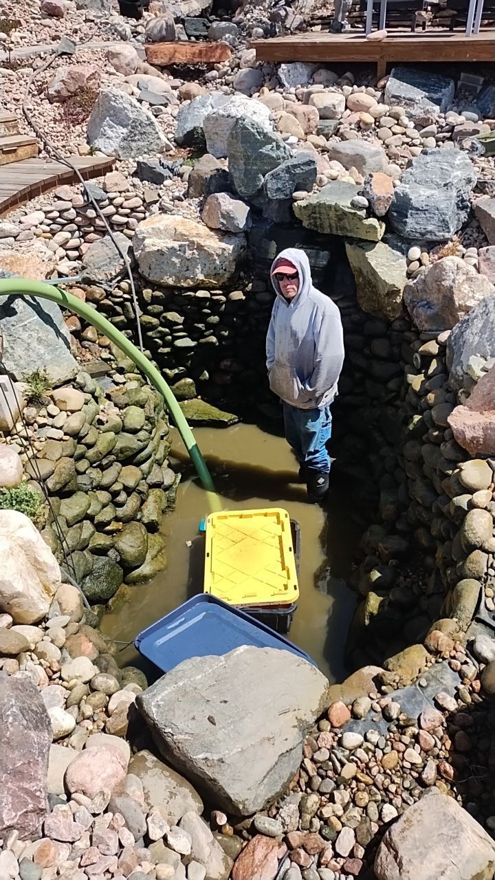 A man in a hooded jacket is standing in a rocky area with a hose.