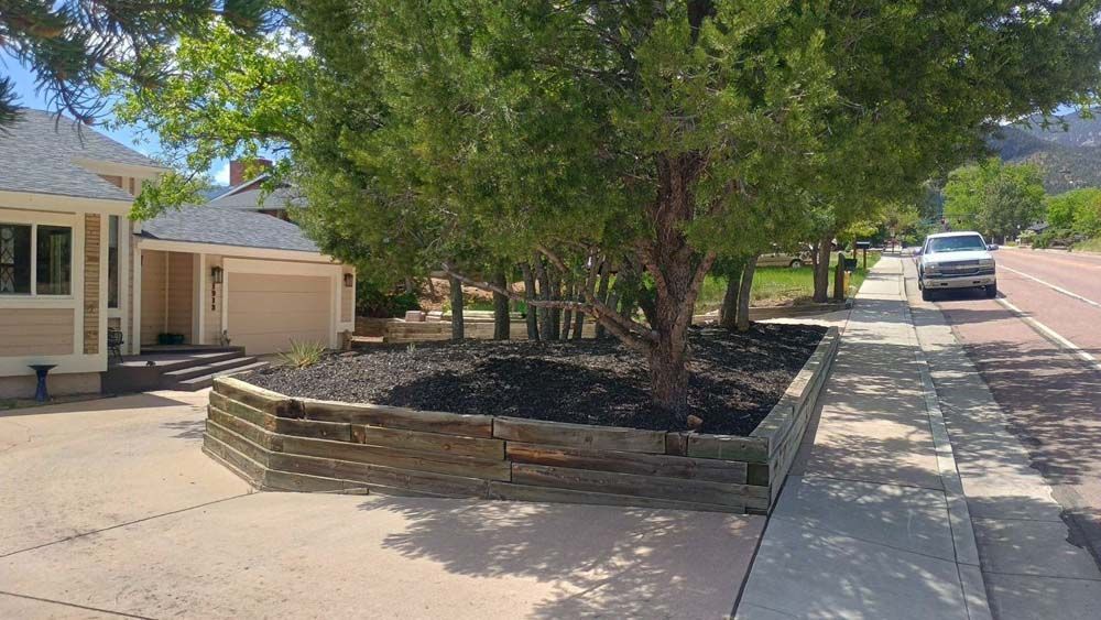 A house with a beige garage and a retaining wall with mulch and trees next to a sidewalk and street.