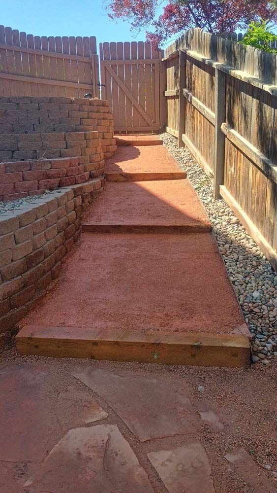 Pathway with steps made of reddish gravel, bordered by wooden fence and retaining walls, leading to a gate.