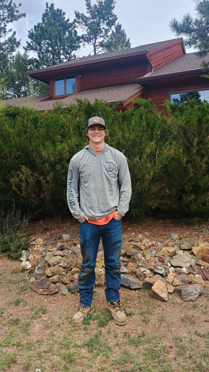 Man in gray hoodie and jeans standing in front of a house, hands in pockets.