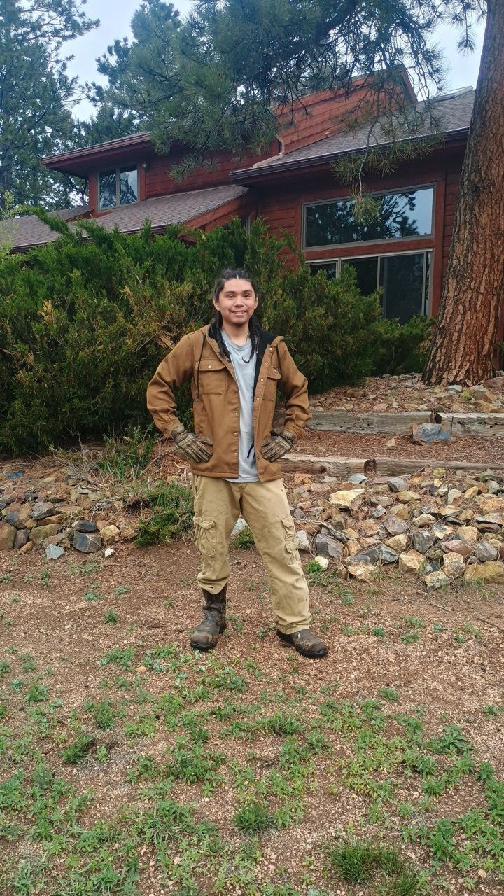 Man in brown jacket and khaki pants stands in front of a house with large windows.