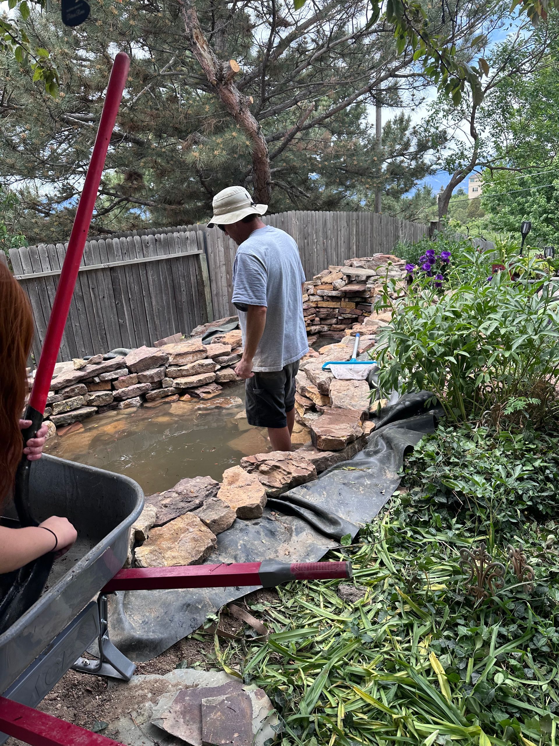 A man is standing in a pond while a woman pushes a wheelbarrow.