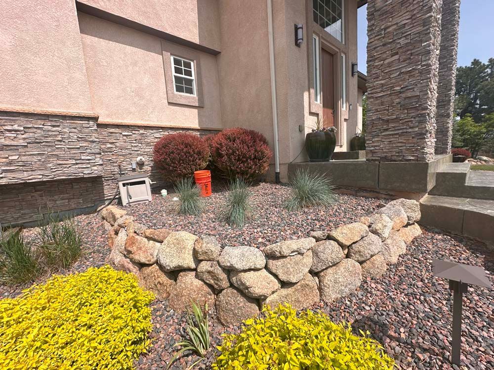 Stone-walled flower bed with yellow and green plants, red mulch, and brown bushes in front of a tan house.